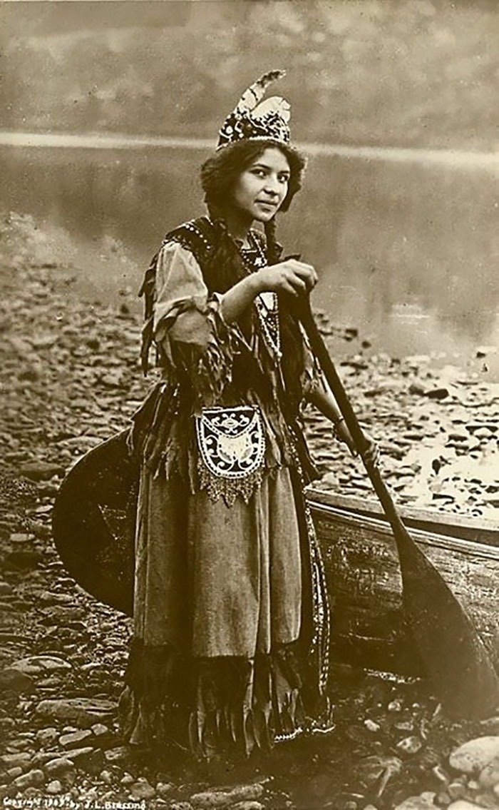 Native American woman in traditional clothing holding a paddle by a riverbank in a historic monochrome photo.