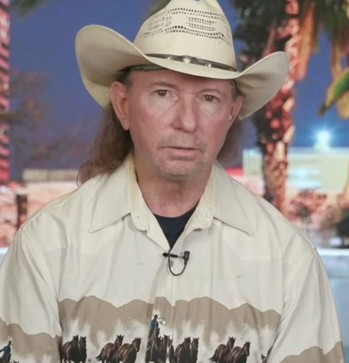 Cowboy in patterned shirt and hat, serious expression, indoor setting with palm decor.