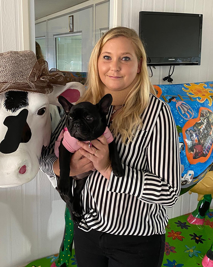 Woman holding a small black dog, standing next to a colorful cow statue, related to Virginia Giuffre’s tragic final note. Woman holding a small black dog, standing next to a colorful cow statue, related to Virginia Giuffre’s tragic final note.