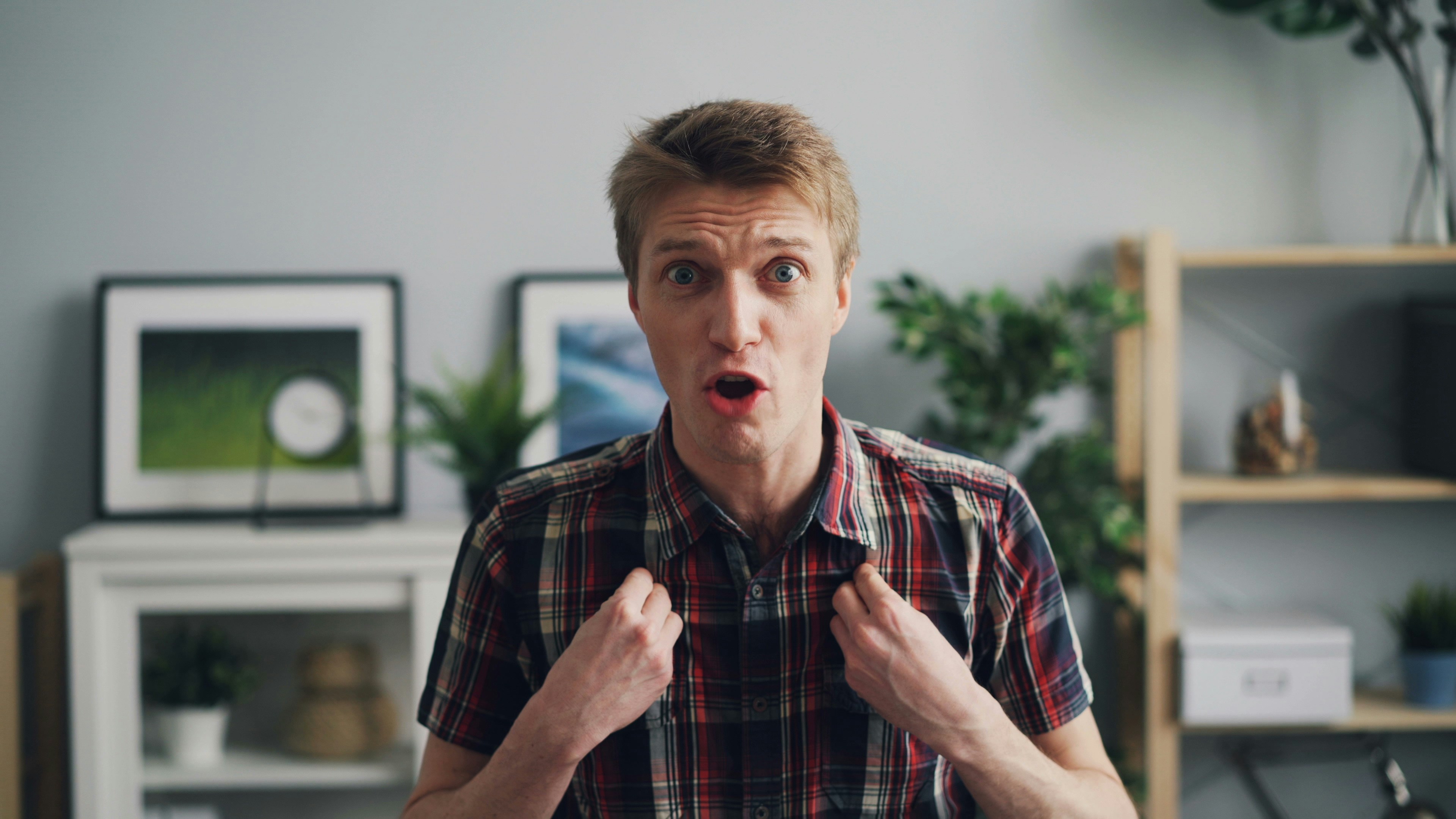 A surprised man in a plaid shirt gestures emotionally, set against a home office background with plants and shelves.