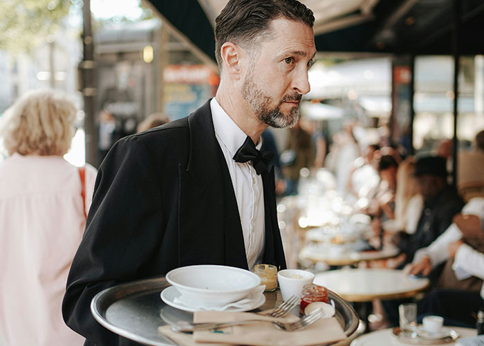Waiter carrying a tray in a busy cafe, looking serious, possibly due to a low tip.