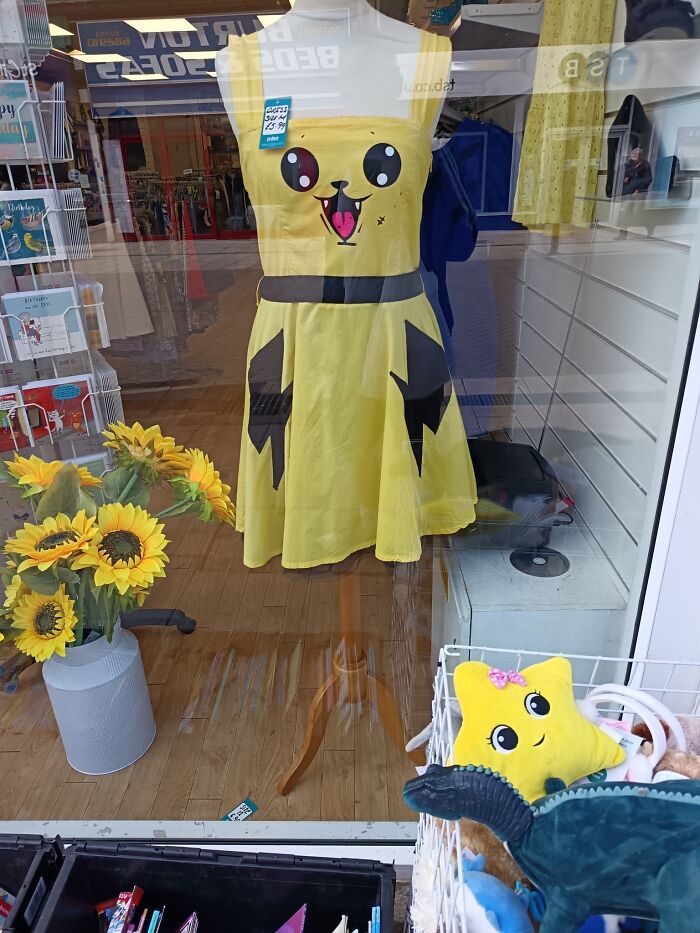 Yellow dress with a playful cartoon face in a secondhand shop window, surrounded by sunflowers and plush toys.