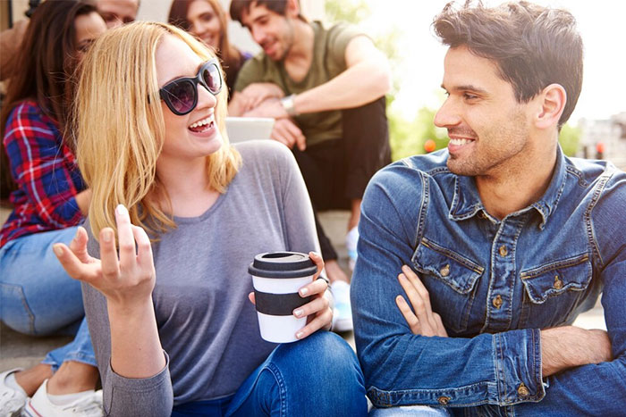 Friends sitting together outdoors, woman in sunglasses holding a coffee cup, smiling, and chatting with a man in a denim shirt.