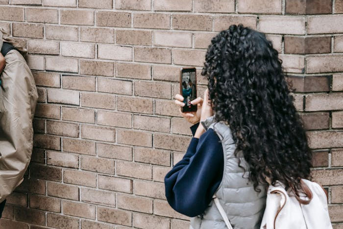 Person with curly hair taking a photo on their phone against a brick wall backdrop, capturing a moment.
