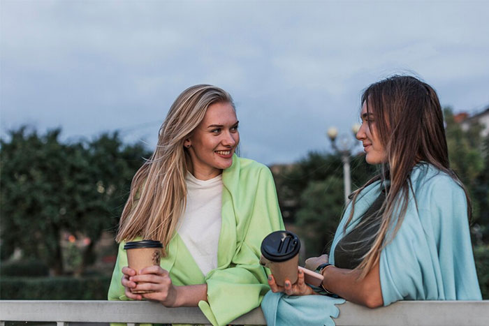 Two friends chatting on a bridge, wrapped in blankets, enjoying coffee; depicting a friendship moment.