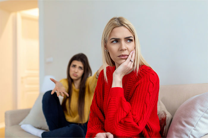 Two women on a couch, one in red sweater looks away thoughtfully while the other gestures in frustration, highlighting friendship tension.
