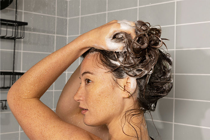 Woman washing her hair with shampoo in a tiled shower.