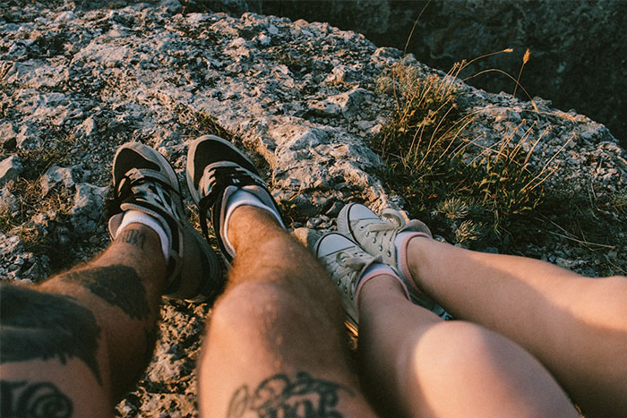 Two friends with tattoos sitting on rocky cliff, shoes touching, representing friendship ending stories theme.