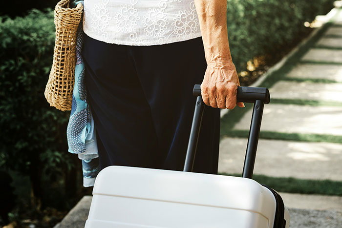 Elderly woman with suitcase, standing on a path, carrying a woven bag. Elderly woman with suitcase, standing on a path, carrying a woven bag.