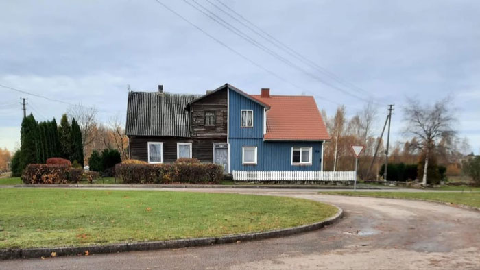Half wooden, half blue-paneled house with red roof on a corner lot surrounded by grass and bushes, unusual real estate property.