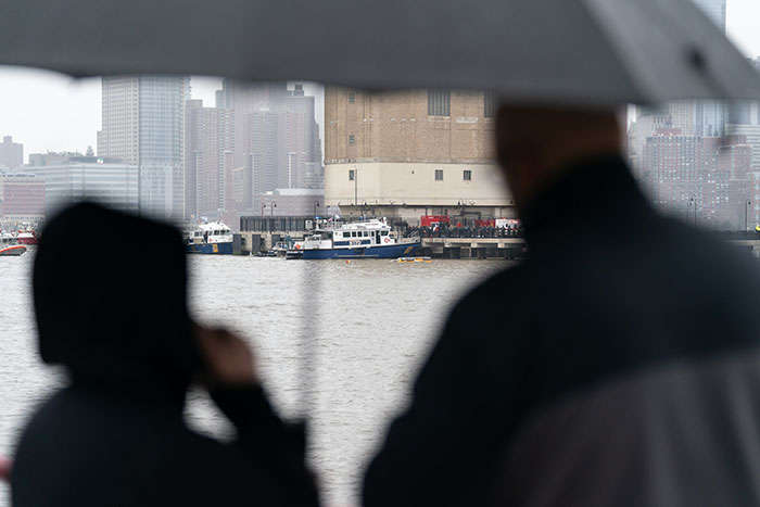 Silhouettes of onlookers near NYC helicopter crash site by the river under an umbrella, with rescue boats in the background.