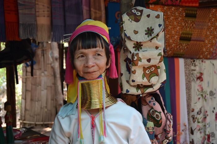 A woman in traditional attire with neck rings in a market setting, surrounded by colorful textiles and bags.