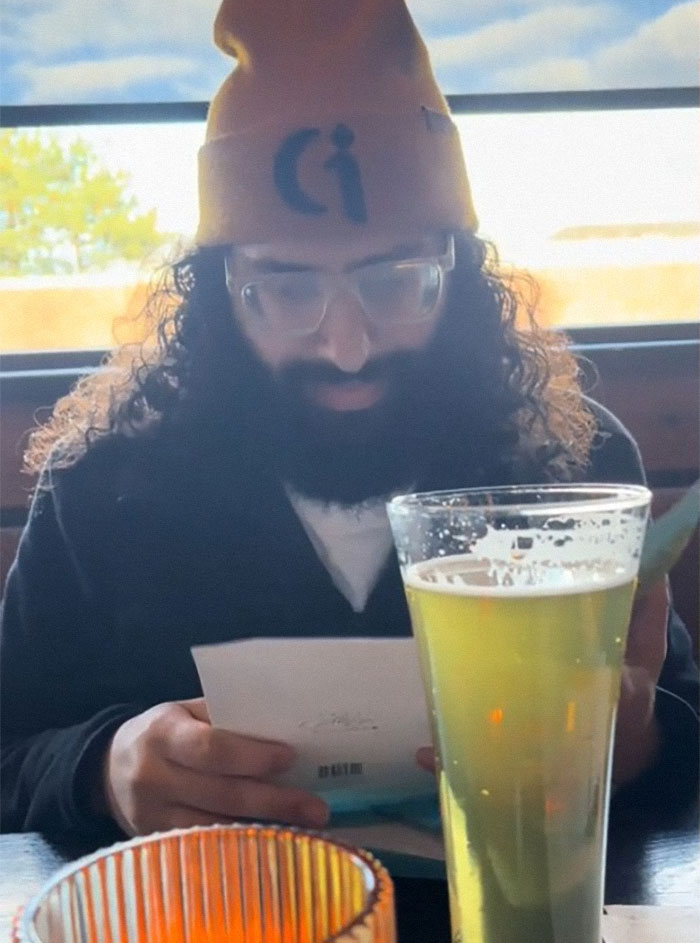 Man reading surprising pregnancy results at a table, wearing glasses and a yellow beanie.