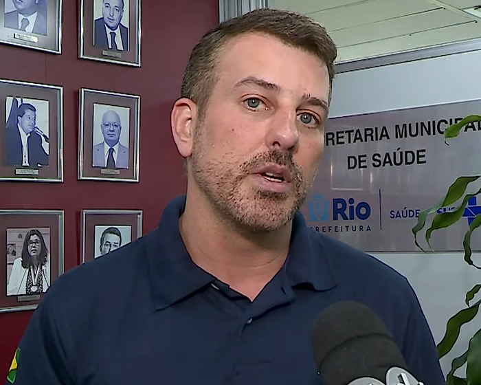 Man speaking at health department office, wearing a navy polo, featured against a backdrop of framed portraits.