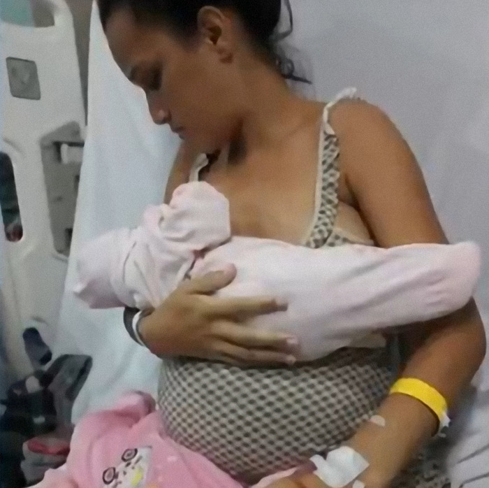Pregnant woman holding one baby in hospital bed, wearing a patterned dress and yellow wristband.
