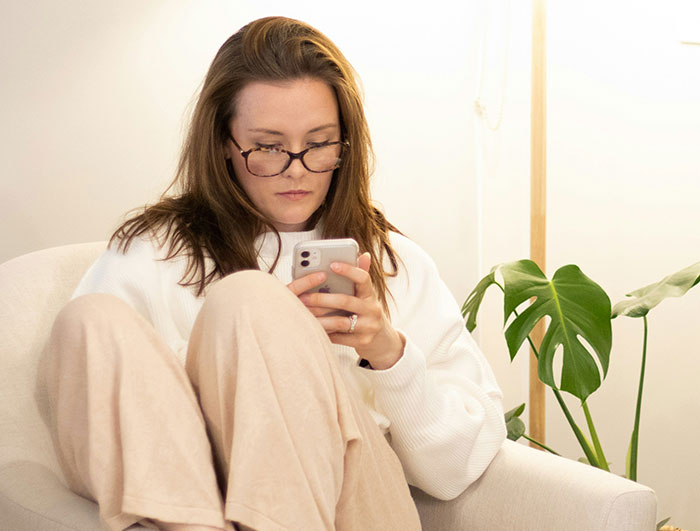 Woman in glasses looking at phone, wearing a white sweater, seated near a plant, receiving text about weight comment.