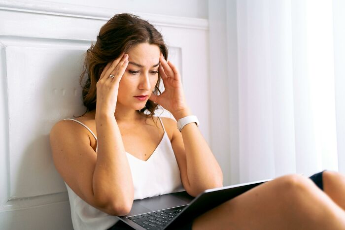 A thoughtful woman sits near a wall, holding her head, with a laptop on her lap, regarding a sister-in-law housing issue.