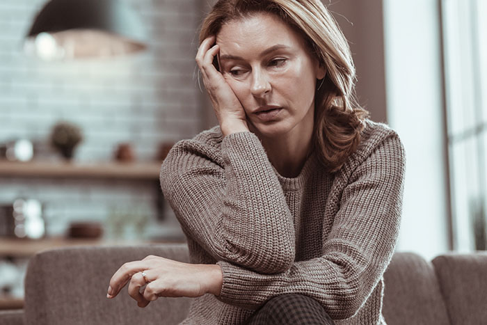 Woman looking pensive, sitting with her hand on her face, in a living room setting.