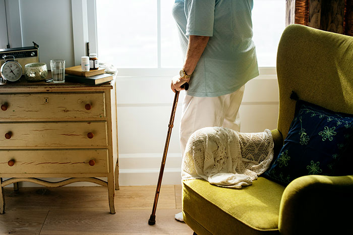 Elderly person with cane beside a chair and dresser in brightly lit room.