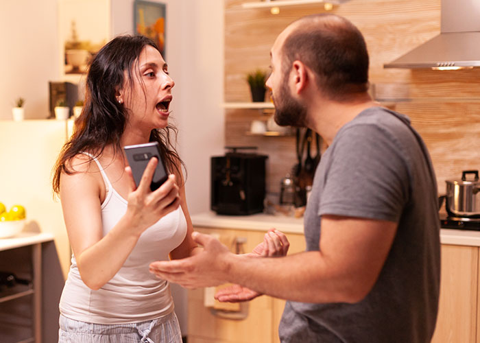 Woman holding phone arguing with husband in kitchen over kids' schedule. Woman holding phone arguing with husband in kitchen over kids' schedule.