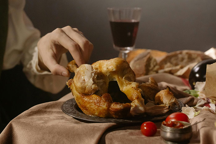 A hand reaching for a roast chicken on a dining table with wine, bread, and tomatoes, highlighting family food dynamics.