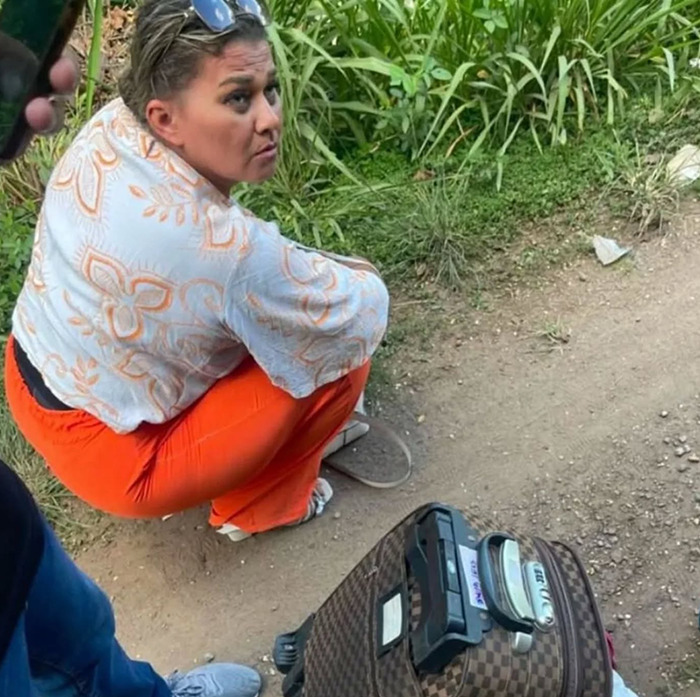 Woman with suitcase on path near greenery, wearing orange pants and patterned top, associated with poisoned Easter eggs incident. Woman with suitcase on path near greenery, wearing orange pants and patterned top, associated with poisoned Easter eggs incident.