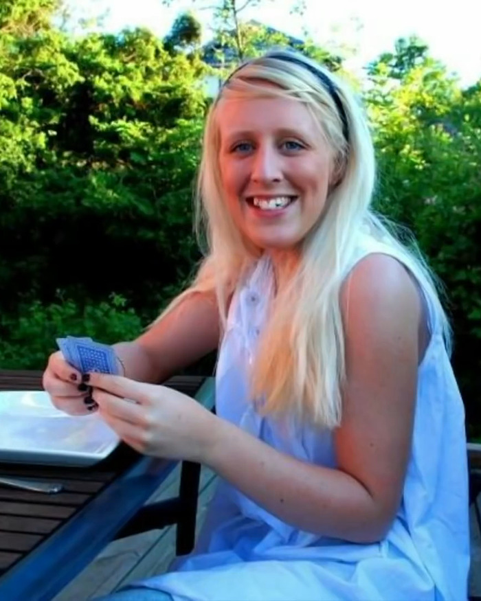 A woman with long blonde hair sitting outside, smiling and holding playing cards.