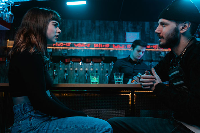 A woman and a man in deep conversation at a bar, with another man in the background serving drinks.