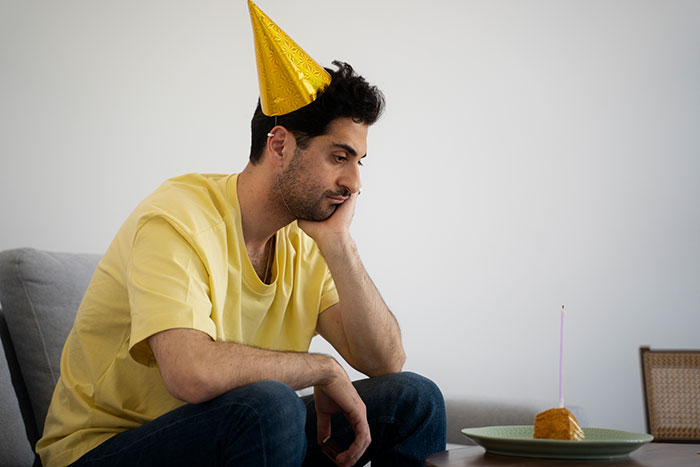 Man in yellow shirt and party hat looking unhappy at a small cake with candle, related to birthday dinner celebration.
