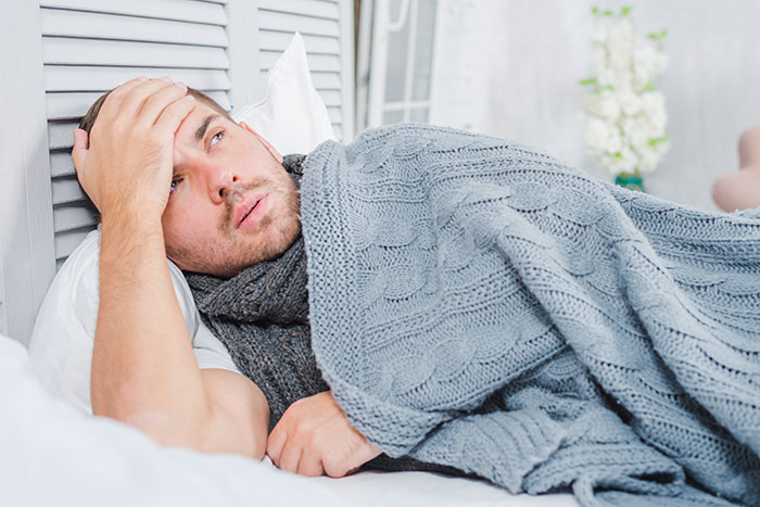 Man lying in bed under a gray blanket, holding his forehead, appearing to be in pain.
