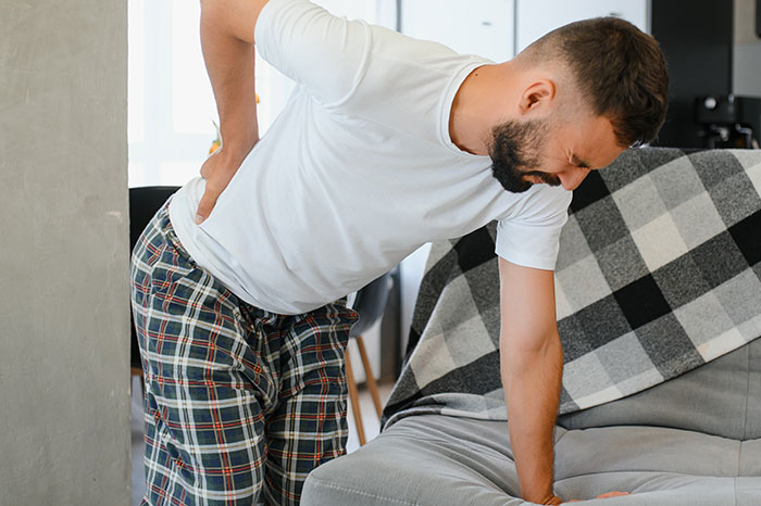 Man in pain holding his back, wearing white t-shirt and plaid pants, near a sofa.