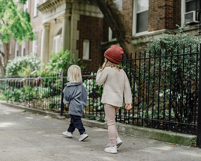 Two toddlers walking on a sidewalk, one wearing a red hat, near a brick building, SEO keywords: toddlers wandering. Two toddlers walking on a sidewalk, one wearing a red hat, near a brick building, SEO keywords: toddlers wandering.