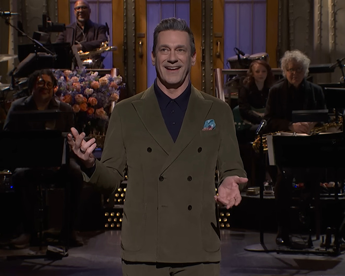 Jon Hamm in a suit, speaking on a stage, with musicians in the background.