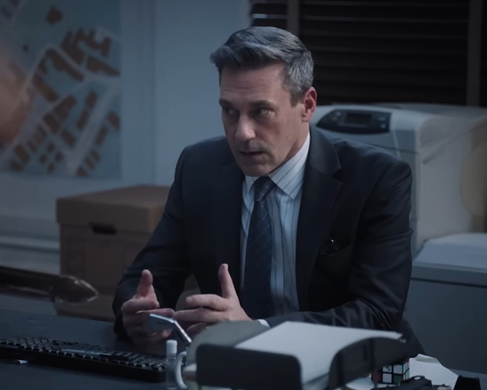 Man in a suit sits at a desk in an office setting, discussing Jon Hamm&rsquo;s university past.