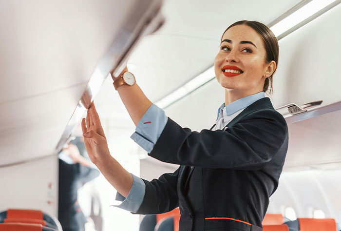 Flight attendant closing overhead bin, smiling, inside airplane cabin.