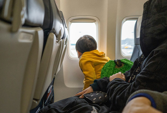 Child in colorful clothes looking out airplane window, seated next to a resting passenger.