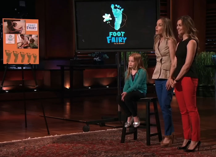 Three women presenting the Foot Fairy product on Shark Tank stage, one seated and two standing, with product display boards nearby.