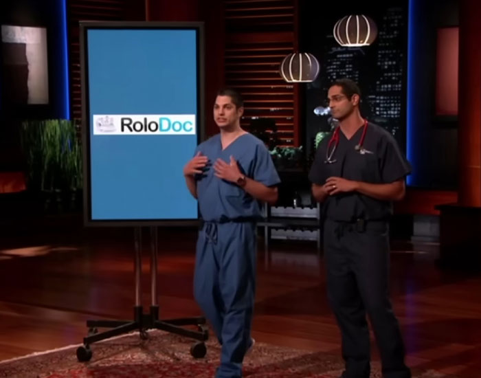 Two men in medical scrubs presenting Shark Tank products with a display screen showing a product logo indoors.