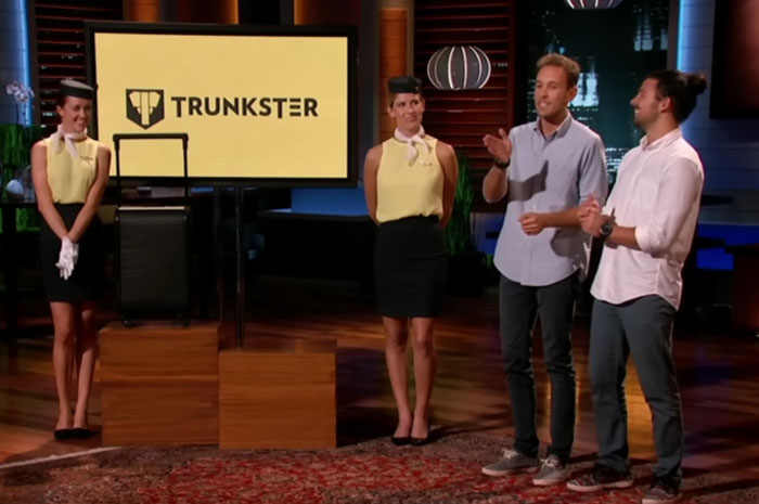 Two men presenting Shark Tank products alongside two women dressed as flight attendants near a screen displaying Trunkster logo.