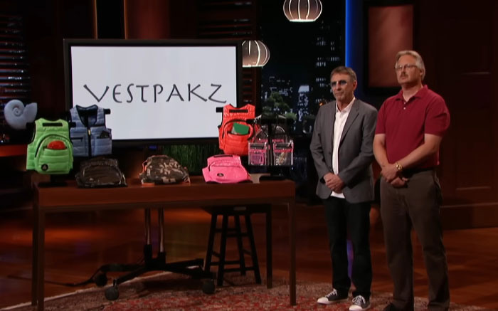 Two presenters stand next to a table displaying Vestpakz products on Shark Tank, a notable Shark Tank product flop.