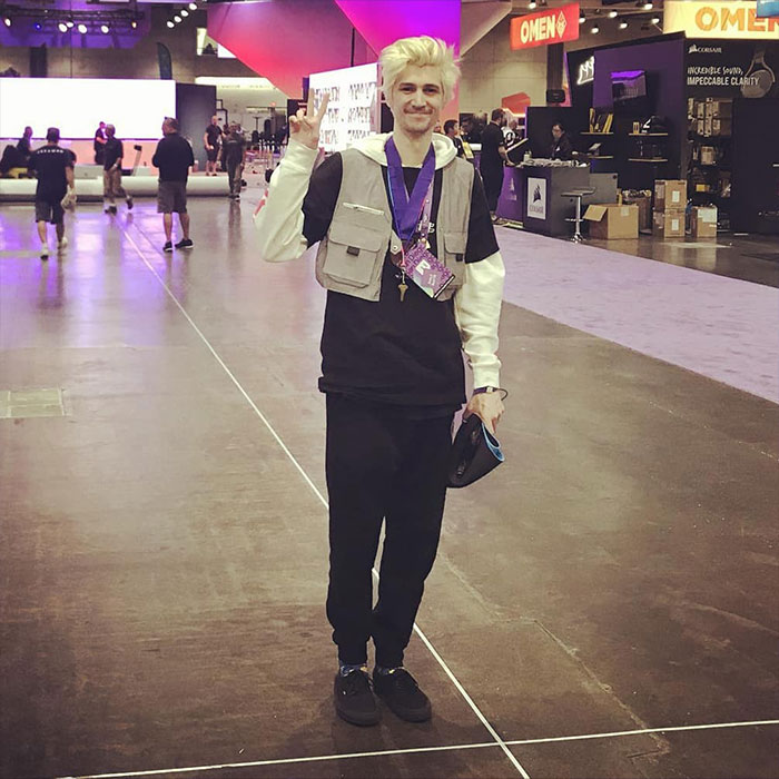 Streamer xQc at a gaming convention, wearing a vest and lanyard, standing on a large showroom floor.