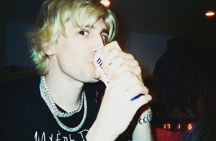 Streamer xQc drinking from a can, wearing a black shirt and silver chain, with dim lighting in the background.
