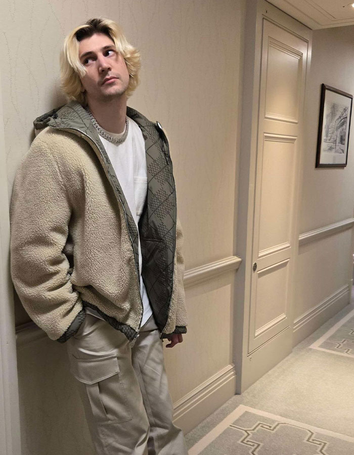 Streamer xQc standing in a hallway wearing a tan jacket and cargo pants, associated with xQc net worth discussions.