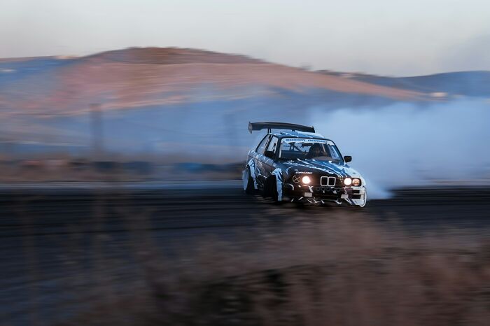 A racing car drifts on a track, smoke trailing behind, illustrating hazardous conditions EMTs and first responders may face.