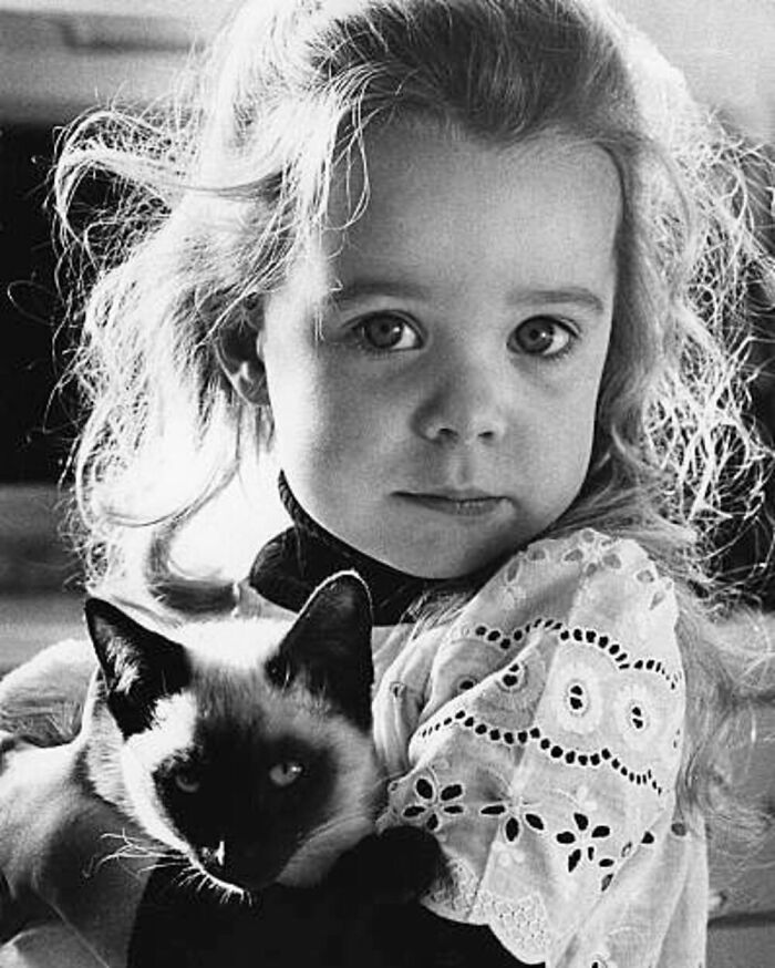 Vintage black and white photo of a young girl lovingly holding a Siamese cat, showcasing the timeless bond between kids and cats.