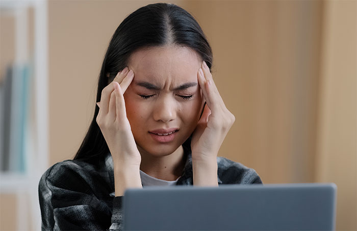 Young woman showing signs of attention seeking with a pained expression while working on a laptop in an office setting.