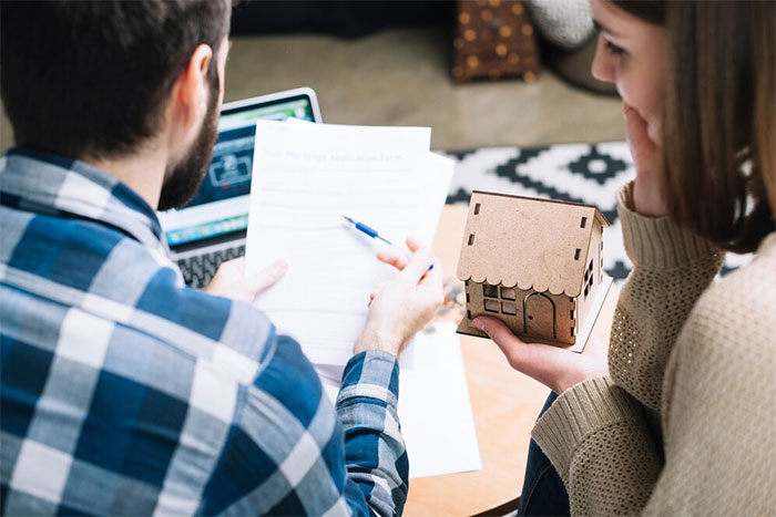 Couple reviewing estate documents while woman holds small wooden house model, reflecting on late father’s estate inheritance. Couple reviewing estate documents while woman holds small wooden house model, reflecting on late father’s estate inheritance.