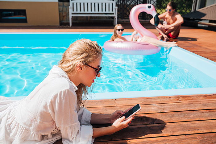 Woman in sunglasses using phone by pool while neighbors&rsquo; relatives enjoy pool with inflatable flamingo nearby.