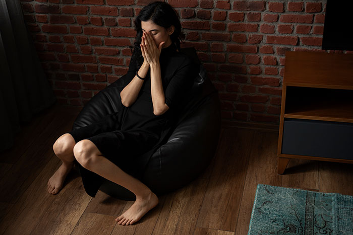 Woman showing signs of being too depressed to babysit, sitting on a black bean bag chair in a dimly lit room.