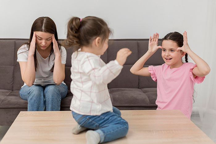 Woman looking overwhelmed and depressed babysitting two energetic children playing on a couch and table indoors.